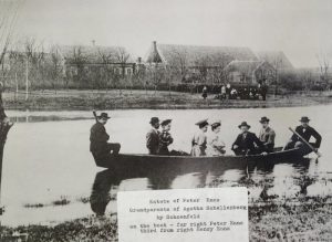 Black and white photograph of five men and three women in a boat. Man at front and man at back have paddles. Behind them is a collection of buildings. They are fancily dressed with suits and hats