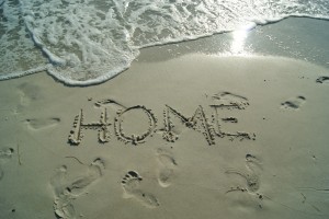 Footprint and the word "home" drawn into beach sand. A wave is getting close.