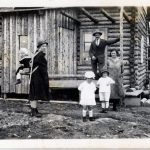 Black and white photograph of a family standing on the porch of their log home. The mother is wearing her child on her back in a type of frame