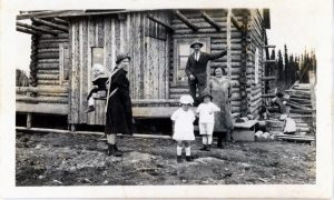 Black and white photograph of a family standing on the porch of their log home. The mother is wearing her child on her back in a type of frame