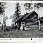 Black and white photograph of the side view of a log cabin in front of a few pine trees