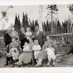 Black and white photograph. This is a group portrait of an extended family. The front row of women and babies are sitting and the men stand behind. Two men are holding babies. The men are wearing suits and ties. Behind them is a forest of pine trees