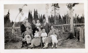 Black and white photograph. This is a group portrait of an extended family. The front row of women and babies are sitting and the men stand behind. Two men are holding babies. The men are wearing suits and ties. Behind them is a forest of pine trees